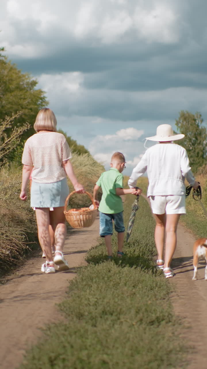Back view of family walking on dirt road surrounded by lush greenery during retreat, carrying picnic basket while aunty holds umbrella and dog on leash, under partly cloudy sky