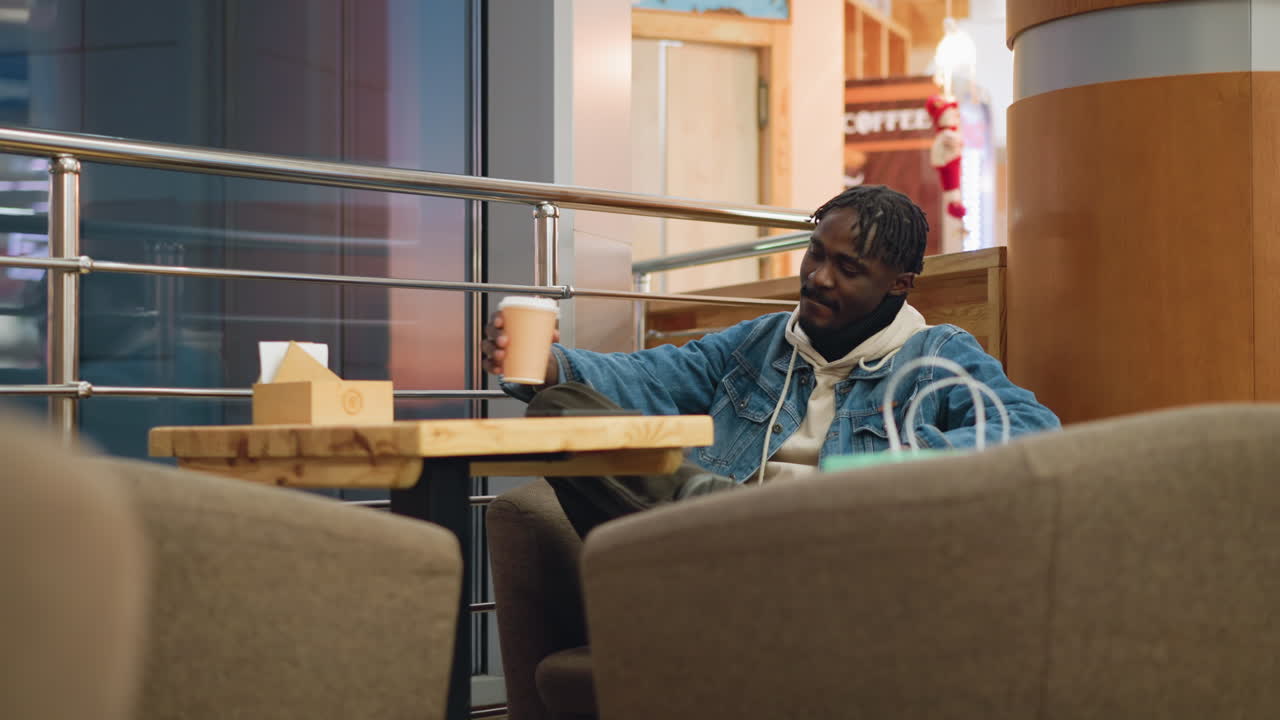 Guy wearing denim jacket and hoodie sits alone in cozy caf lounge, holding coffee cup with relaxed posture, surrounded by soft chairs, wooden table, modern railings, shopping bags beside him