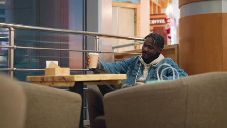 guy wearing denim jacket and hoodie sits alone in cozy caf lounge, holding coffee cup with relaxed posture, surrounded by soft chairs, wooden table, modern railings, shopping bags beside him