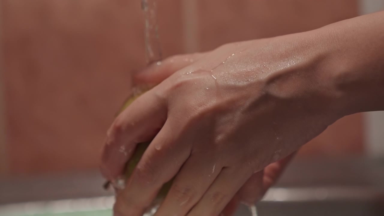 Primo piano di mani che lavano una mela verde sotto l'acqua corrente. Le mani strofinano e ruotano il frutto con goccioline che schizzano. L'area del lavello presenta piastrelle sfocate sullo sfondo, mentre la messa a fuoco rimane stabile.