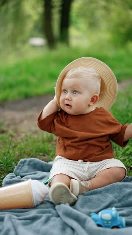 adorable toddler boy tries to put on a straw hat but he fails. funny kid having rest in the nature in summer. blurred backdrop. vertical video.
