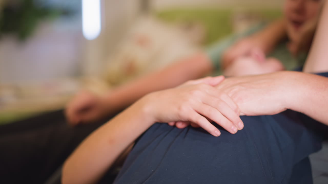 Kvinde hviler på sofa, hånd forsigtigt placeret over torso, hyggelig stue, aftenlys filtrerer gennem vindue, ledsagers arm i baggrunden, nærbillede af hænder, bløde tekstiler, pude, intimt øjeblik af ro