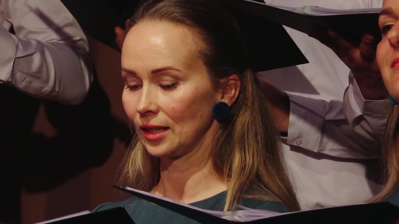 A focused woman sings in a choir, holding sheet music as others around her perform, capturing a moment of vocal expression and concentration.
