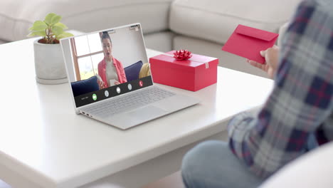 hombre caucásico sosteniendo un sobre rojo usando una computadora portátil con una mujer biracial abriendo un regalo en la pantalla