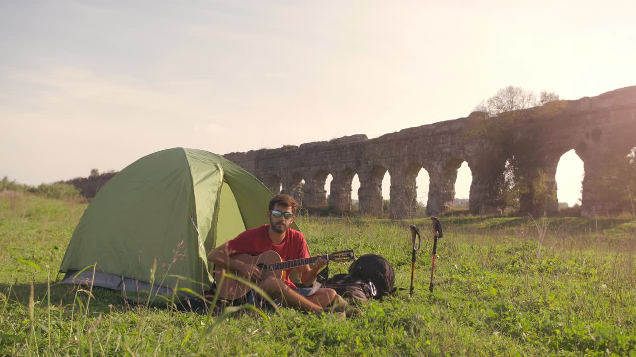 캠핑 텐트 밖에서 기타를 연주하는 젊은 배 타는 남자 로마 수로 앞에 잔디에 앉아 parco degli acquedotti의 공원 유적에서 로마에서 해가 지는 동안 땅 속 느린 동작에 을