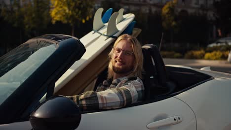 Portrait-of-a-happy-blond-guy-in-glasses-with-a-beard-in-a-checkered-shirt-who-sits-in-a-White-convertible-and-to-the-right-of-him-there-are-two-surfboards-in-the-city