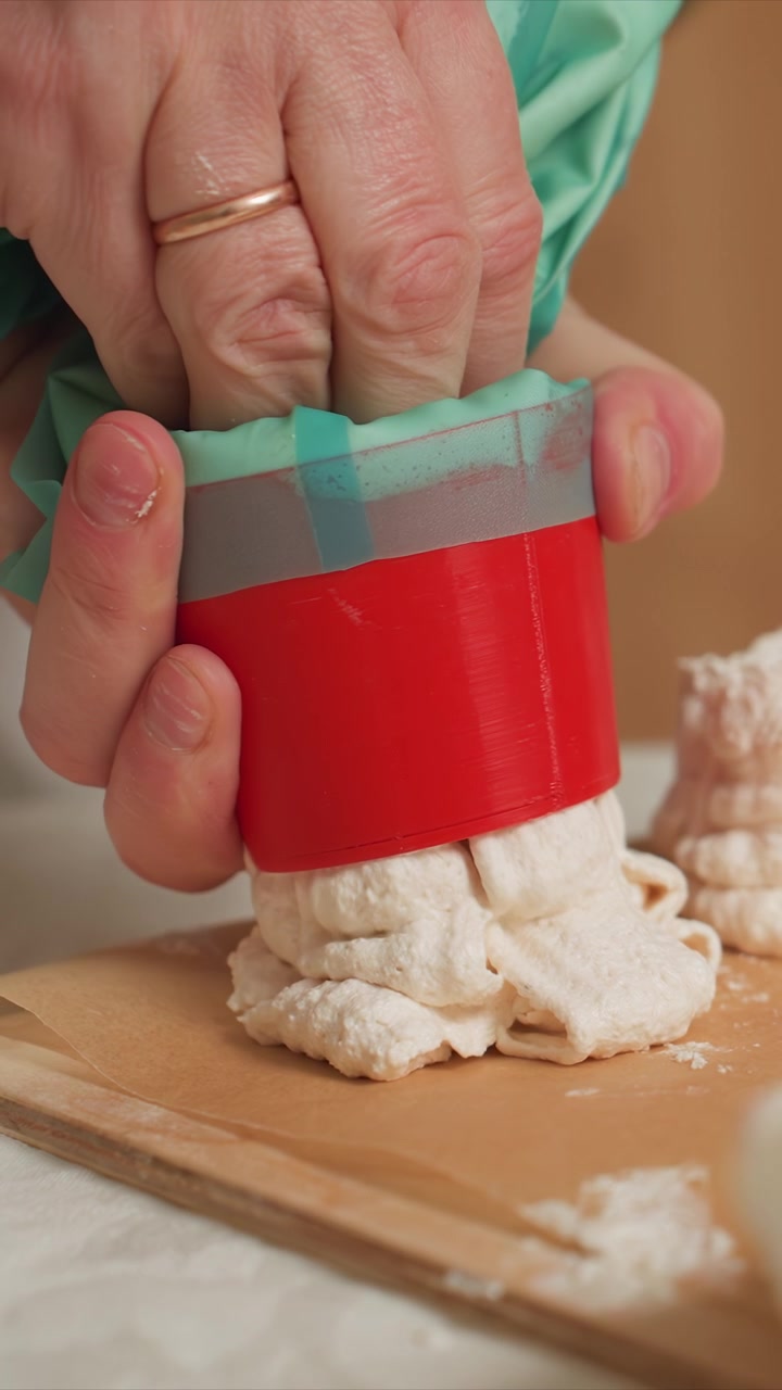 Primo piano di mani che usano uno strumento cilindrico rosso. Il processo prevede la pressione e l'estrusione di una pastella di colore chiaro. Ambientazione interna con toni neutri, concentrandosi unicamente sull'impasto e le mani.