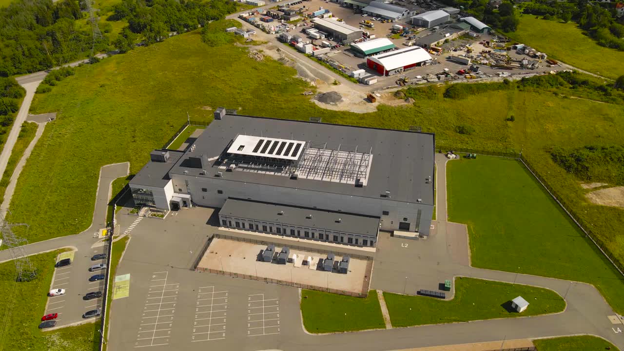 Aerial drone footage orbiting and flying around a large modern gray and white colored data center with cooling system ventilation fans on the rooftops and green grassy countryside around it, servers