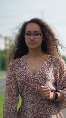 mujer con vestido vintage caminando al aire libre, con la mano sujetando la correa de la bolsa, ajustando el cabello contra el telón de fondo de la vegetación y los peatones lejanos