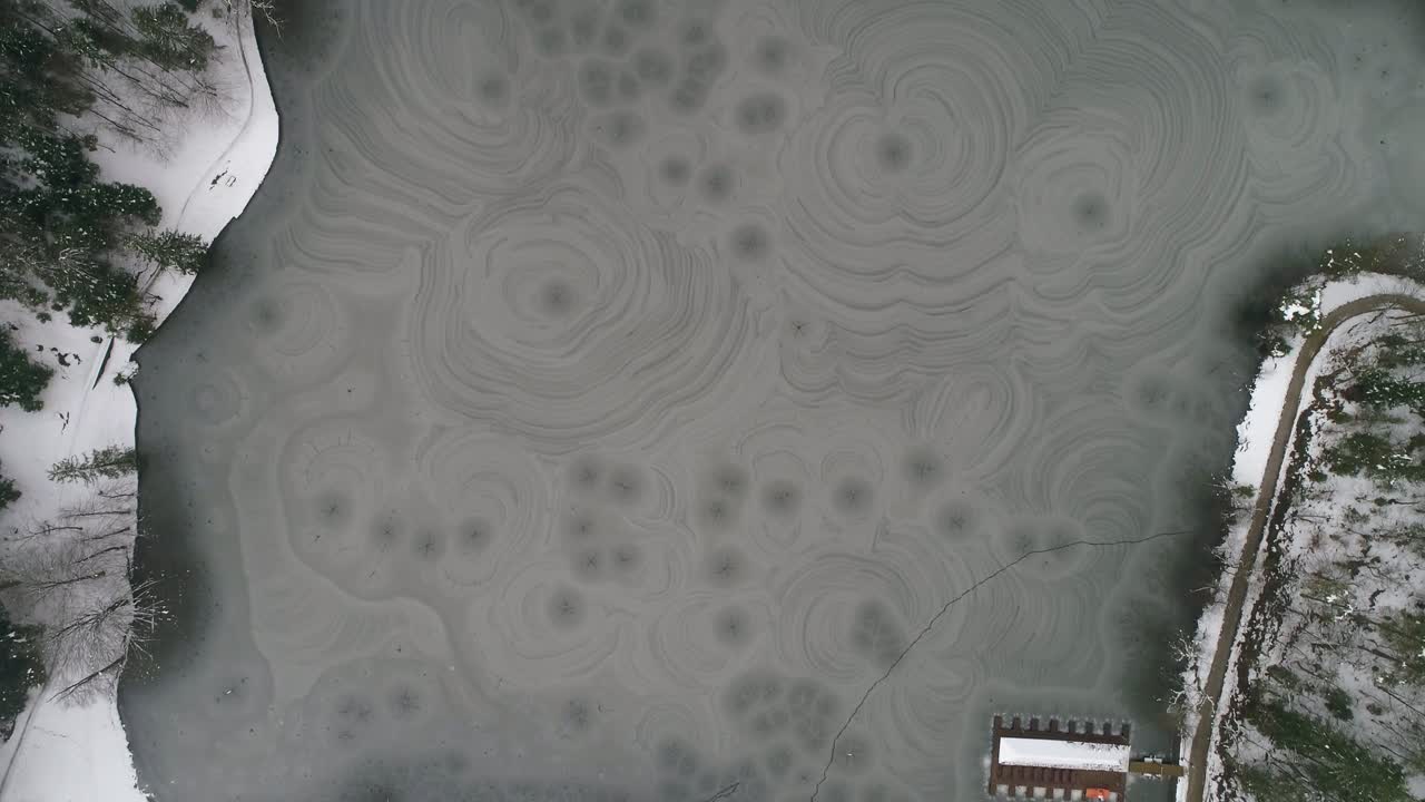 toma aérea de un lago congelado con una pequeña casa en el lago rodeada de bosque en invierno