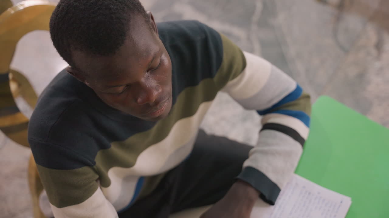 Black man writing love letter at table, young black student scribbling heartfelt note in green notebook, sitting on wooden chair, pen pauses, nervous smile, preparing date surprise, warm soft light