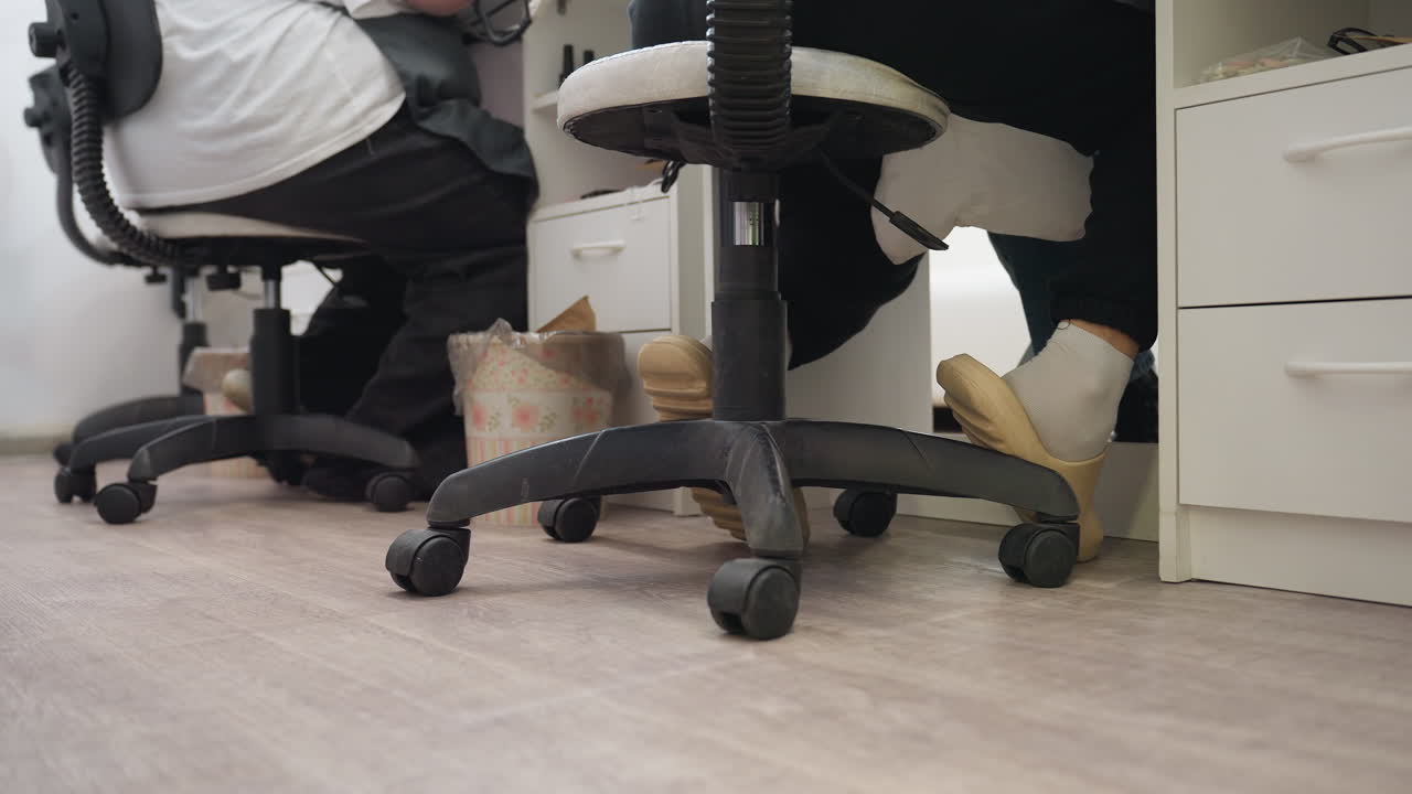 Close-up view of salon floor showing black wheeled chair, technician's feet in beige slippers with white socks, positioned beside white cupboard with drawers in clean, organized workspace