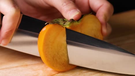 detailed view of hands skillfully slicing a persimmon with a sharp knife on a wooden cutting board.