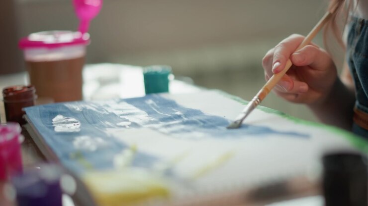 Hand holding brush applies blue paint to paper or canvas. Various colors of paints are visible in jars around workspace suggesting art creation process