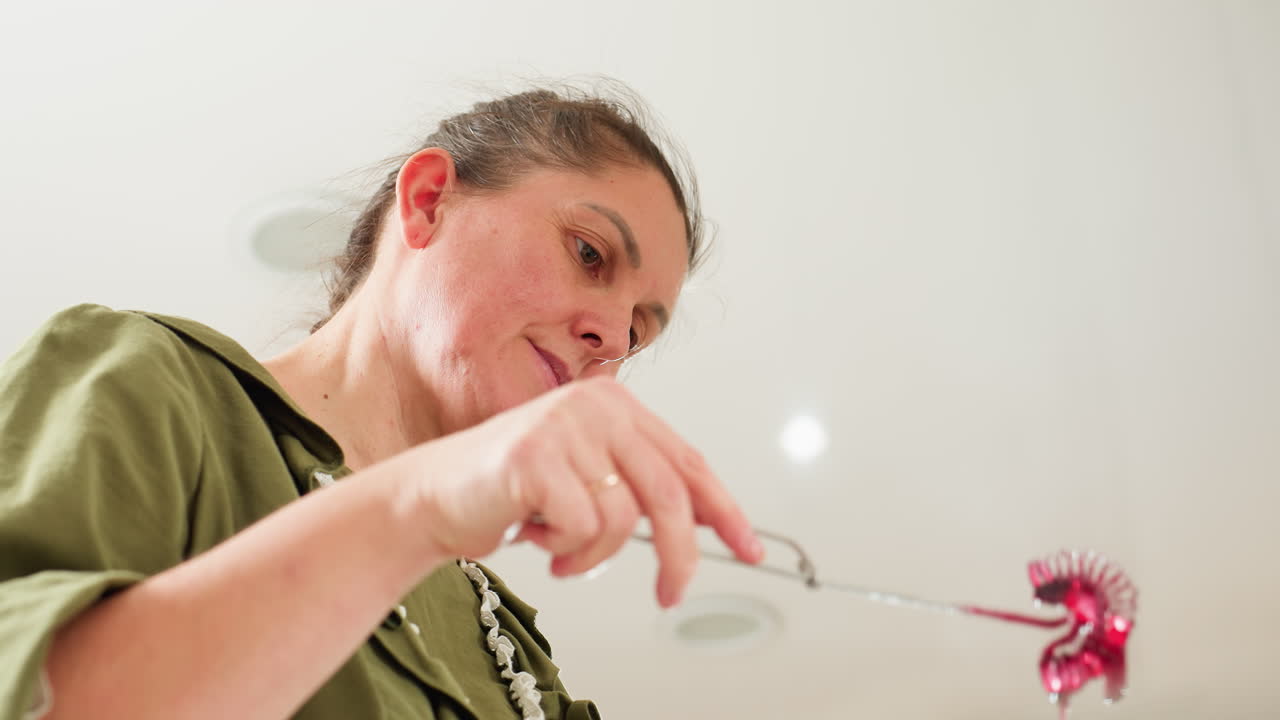 Woman in green blouse examines iron mixer lifted above pot of red soup in bright home kitchen as some drops fall back, showcasing careful cooking process with focus and attention