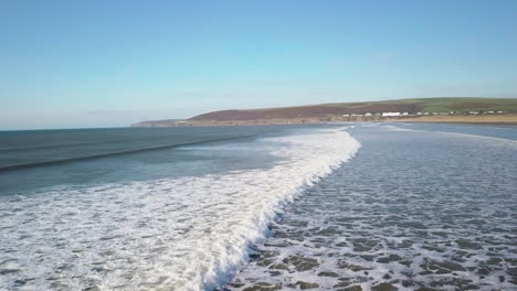 Vuela-Sobre-Las-Olas-Que-Salpican-La-Costa-Bajo-El-Cielo-Azul-De-La-Playa-De-Saunton-Sands,-En-North-Devon,-Inglaterra