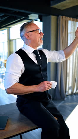 mature businessman wearing official clothes sits on the desk drinking coffee. man is raising his phone trying to catch the mobile connection. vertical video.