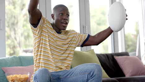 Feliz-Hombre-Afroamericano-Sentado-En-Un-Sofá-Viendo-Un-Partido-De-Rugby-En-Casa,-Cámara-Lenta