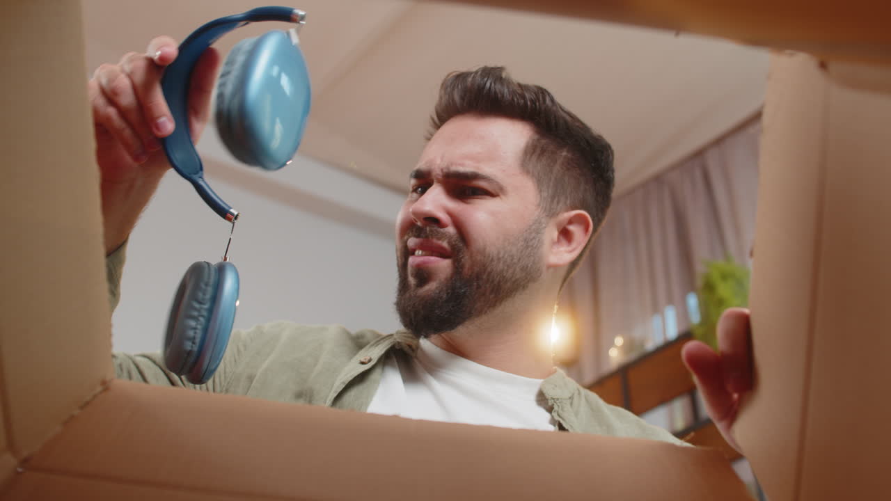 Dissatisfied annoyed male open cardboard box receive damaged broken wireless headphones at home
