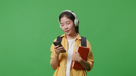 smiling asian woman student with a backpack and some books wearing headphones and using a smartphone while standing in the green screen background studio