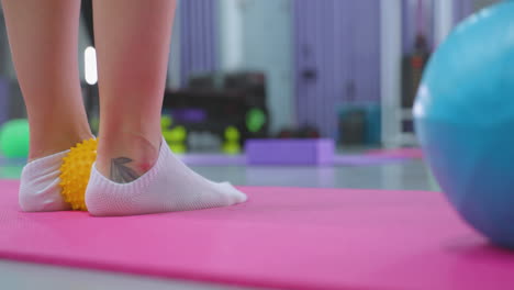 primer plano de una persona levantando ligeramente la pierna en una alfombra rosa con una pelota azul cerca, con equipos de gimnasia y luces parpadeantes vibrantes en el fondo, centrado en la terapia del pie y el ejercicio de equilibrio