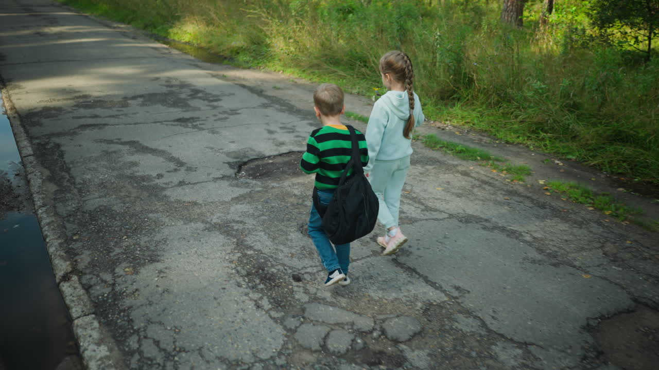 Rear view of little girl holding flower in hand while walking beside her brother on worn asphalt road with visible cracks and potholes, surrounded by grass and forest on sunny peaceful day