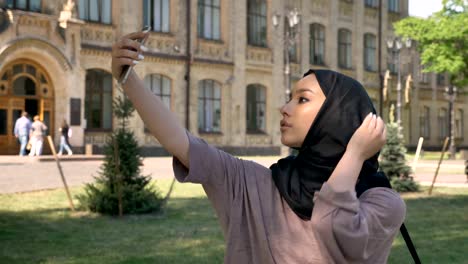 Young beautiful muslim girl in hijab is making selfie on her smartphone in daytime in summer, building on background, religiuos concept, communication concept