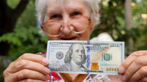 old woman in eyeglasses showing one hundred dollar bill into camera and smiling outdoor. happy grandmother holding foreign currency outside. money concept. close up slow motion