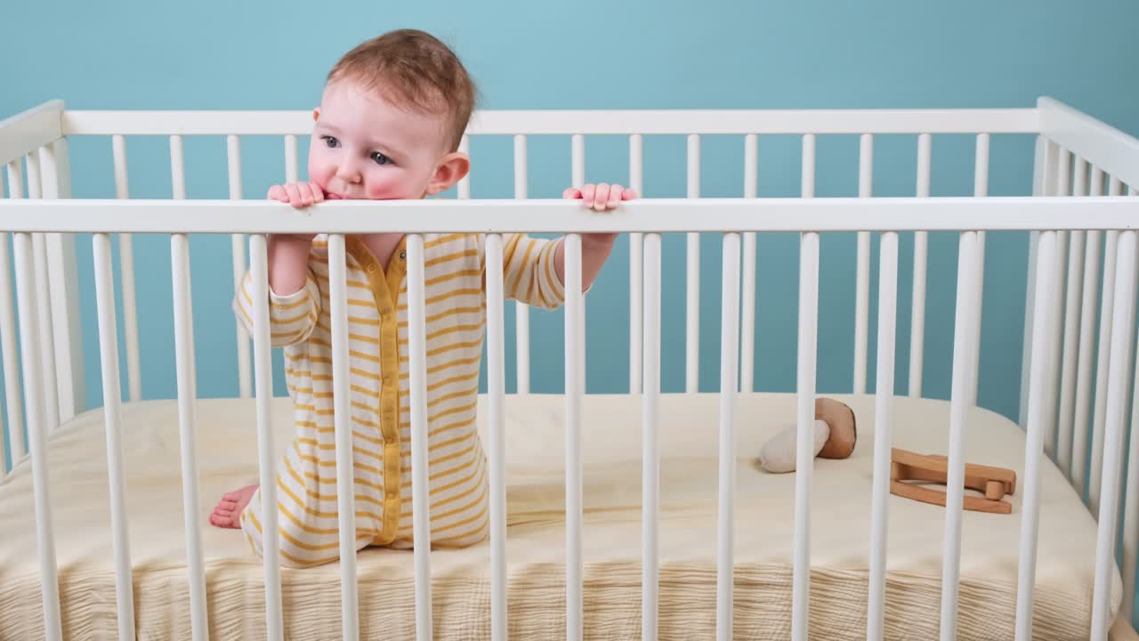 Bambino piccolo triste nella culla che rosicchia le sbarre di legno, sfondo blu da studio. Un bambino stanco con vestiti gialli