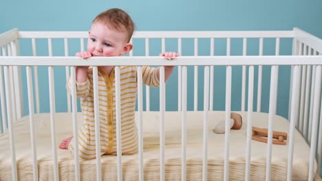 sad toddler baby boy in the crib gnawing a wooden bars, blue studio background. a tired child in yellow pajamas