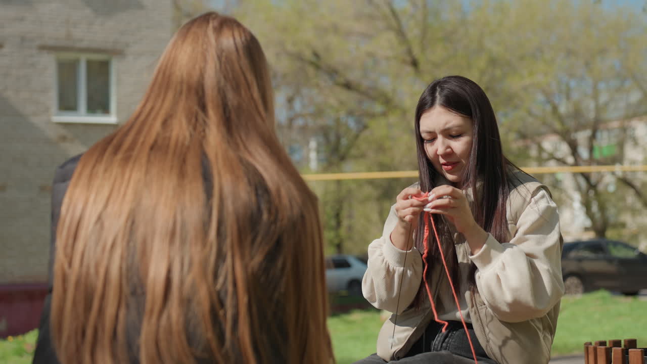 여성이 벤치에 앉은 친구와 함께 야외에서 털실 뜨는 법을 가르치고 있어요. 두 사람은 분홍색 털실과 바늘로 작업을 하면서 팁을 교환하고 있고, 도시 배경에는 건물과 풀이 있으며 협력 학습 분위기가 느껴집니다.