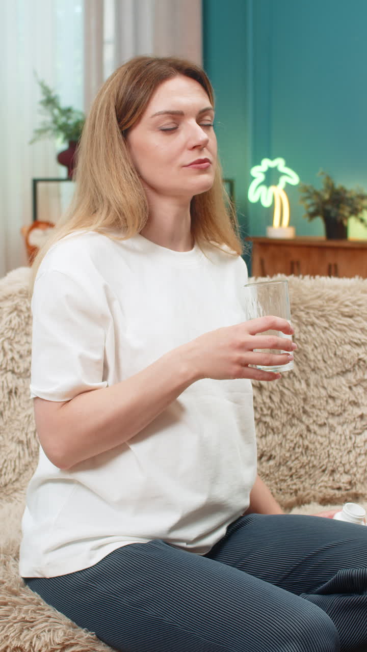 Pregnant woman swallows tablet drinks water sighs relief vitamin treatment routine on sofa at home