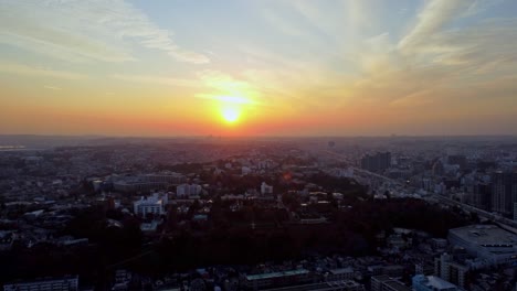 El-Horizonte-De-La-Ciudad-Al-Atardecer-Con-Un-Sol-Radiante-Que-Proyecta-Tonos-Naranja-Y-Azul,-Vista-Aérea