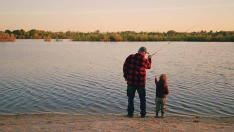 Pesca-Tranquila-En-La-Orilla-Del-Río,-El-Abuelo-Y-El-Nieto-Pequeño-Están-Pescando-Con-Caña-Vista-Trasera