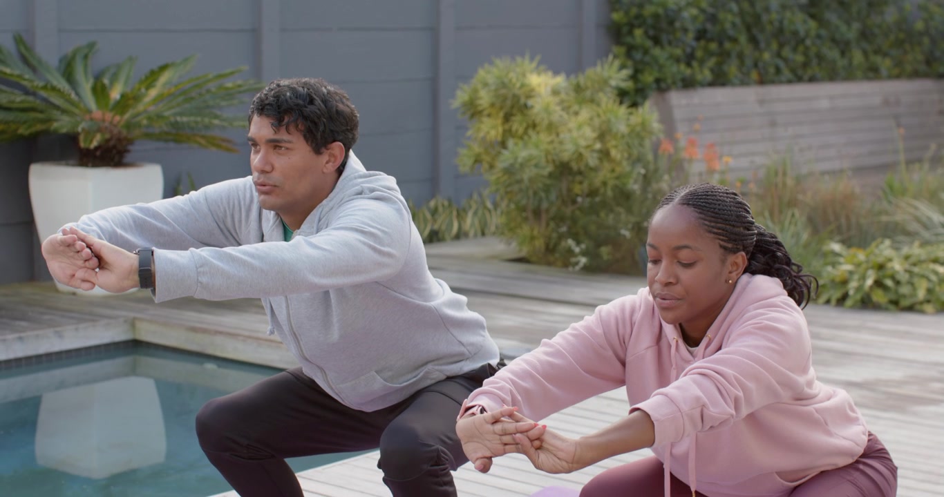 African American couple in hoodies after watch, doing hops, squats for tone on purple mats poolside