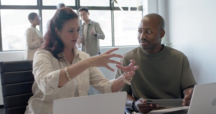 Diverse team meeting at table with monitor showing sales chart initiating woman explaining gestures