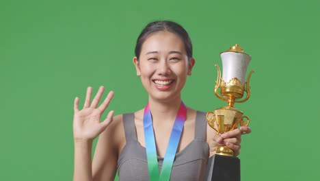 close up of asian woman with a gold medal and trophy waving her hand and smiling to camera as the first winner on green screen background in the studio