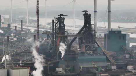 Aerial-medium-shot-of-Tata-steel-mill-with-wind-turbines-in-the-background,-and-one-cutting-through-the-frame