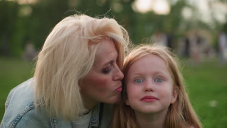 daughter shining eyes while mom gives kiss on cheek in park, joyful closeup showing affection, blurred background of greenery and people, warm golden light highlighting expressions