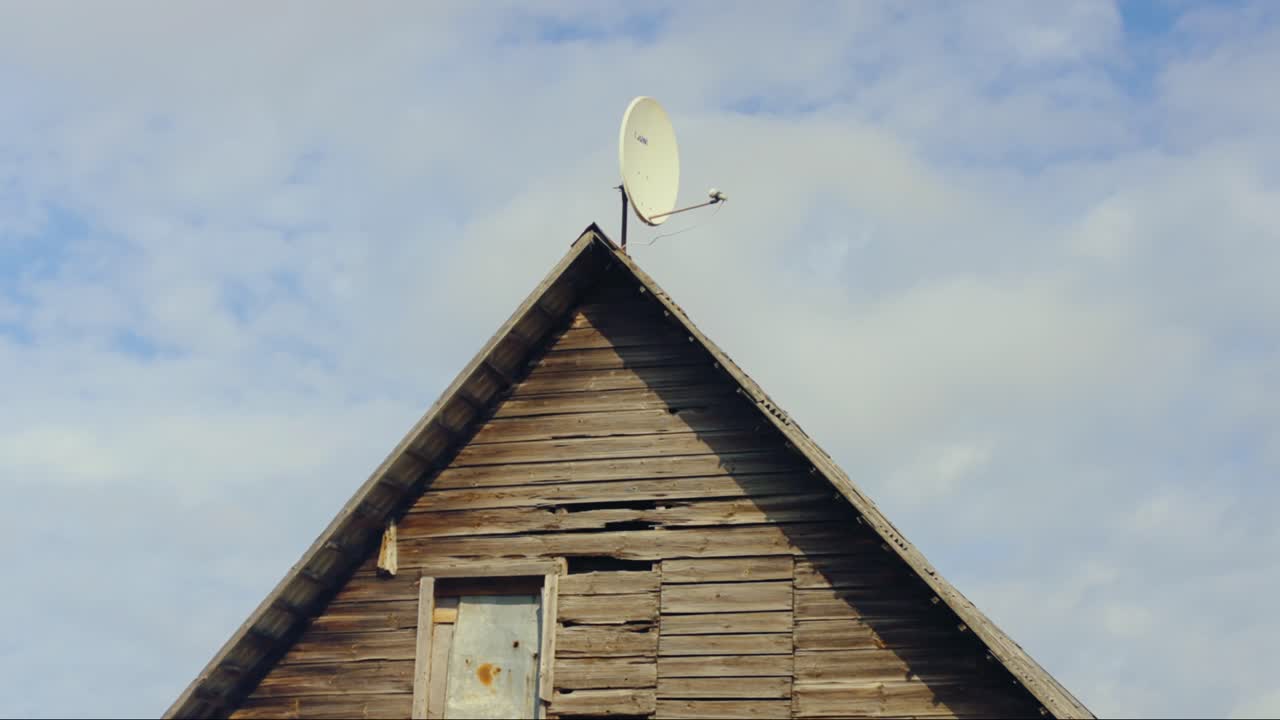 Weathered wooden attic facade featuring a satellite dish mounted at its peak under a blue cloudy sky. Daugavpils, Latvia (Latgale)