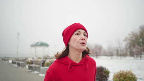 athlete jogging, looking tired along snowy park pathway in red hoodie, surrounded by foggy winter atmosphere with benches, bushes, iron railing by the side and lamp posts behind