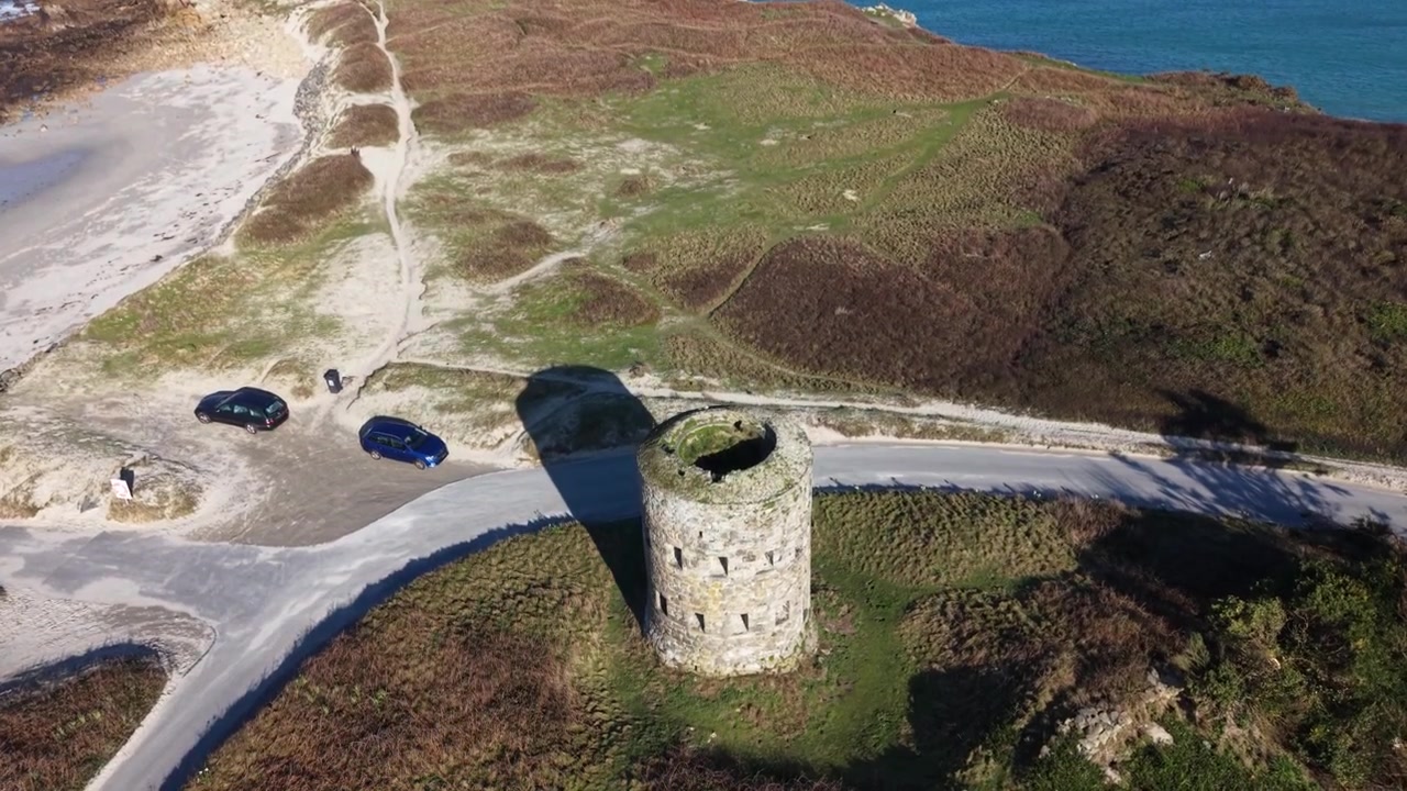 Torre Martello Pembroke Vale Guernsey. Volo alto in avanti sopra la Torre Martello con uccelli in volo e viste sull'intera baia e sulla spiaggia di Lancresse Bay sotto il sole del tardo pomeriggio con cielo azzurro limpido