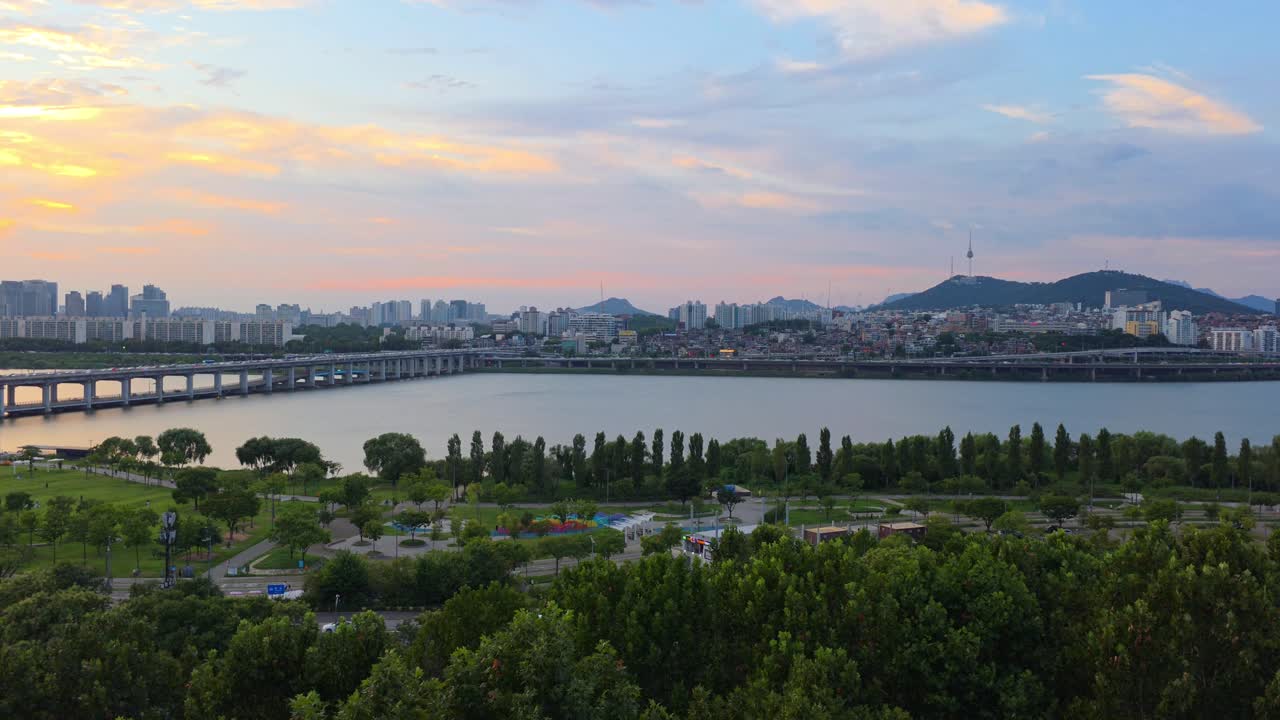 A beautiful wide shot captures the serene blend of nature and city in Seoul, with the lush Hangang Park by the Han River in the foreground and the iconic Namsan Tower against a colorful sunset sky