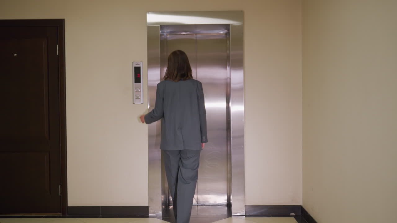 Mujer de negocios en traje gris caminando hacia el ascensor con puertas metálicas cerradas en el piso 3, vista desde atrás en el pasillo de la oficina. La escena refleja la rutina profesional y el ambiente de trabajo.