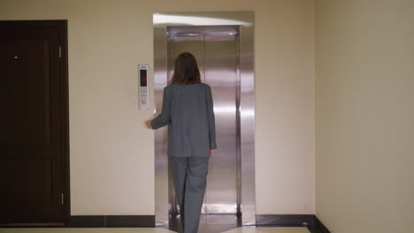 businesswoman in gray suit walking toward elevator with closed metallic doors on floor 3, viewed from behind in office hallway. scene reflects professional routine, workplace environment