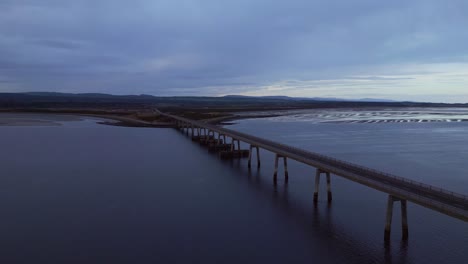 Drone-flight-towards-a-magnificent-Scottish-car-bridge,-embraced-by-the-vast-ocean-and-crashing-waves