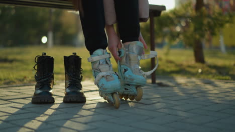 a person wearing a peach jacket and black trousers sits on a park bench, reaching for a rollerblade placed on the ground, preparing to put it on