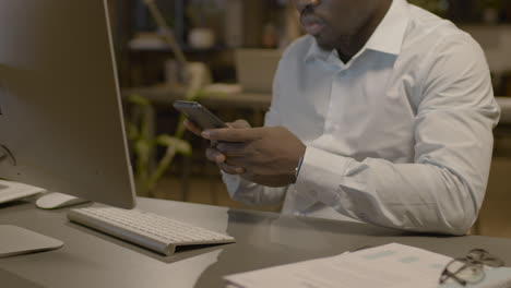 vista de cerca del hombre americano enviando mensajes de texto en el teléfono inteligente sentado en el escritorio en la oficina