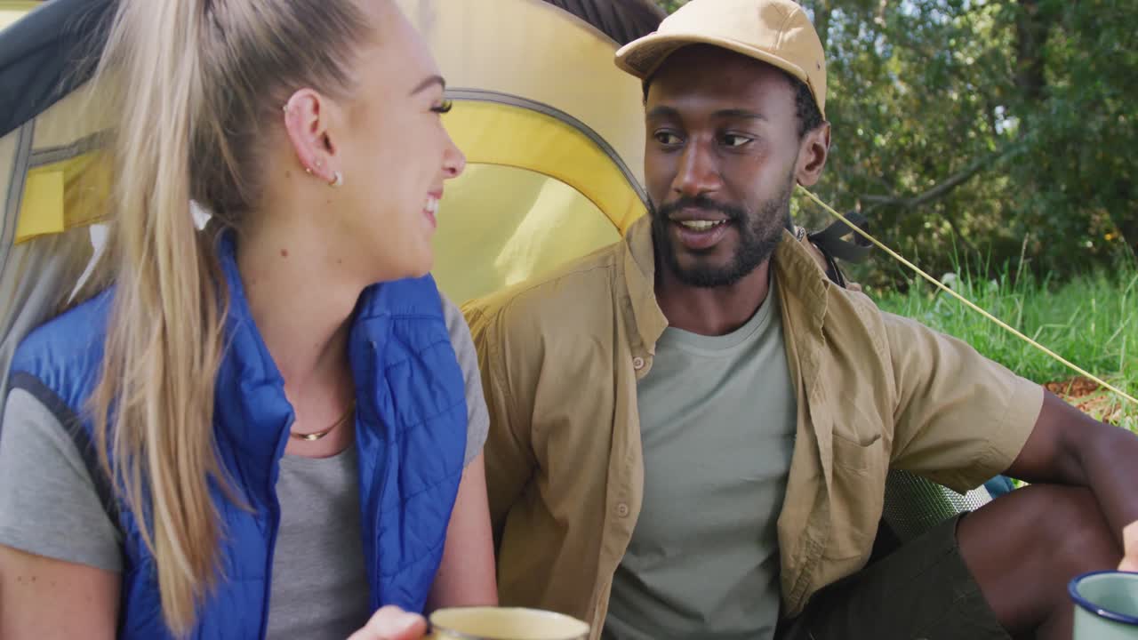 Happy diverse couple camping, drinking tea and talking in park, slow motion