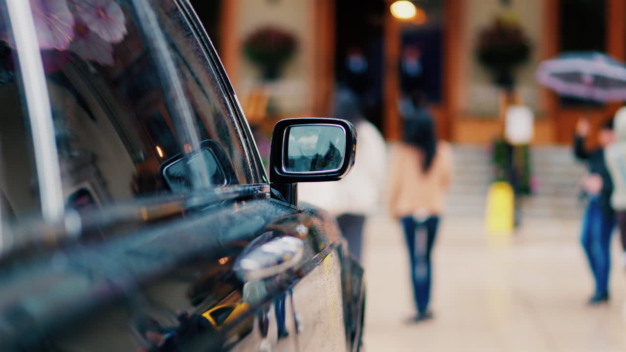 Close up of the mirror of a black car in the rain with people walking on a blurred background